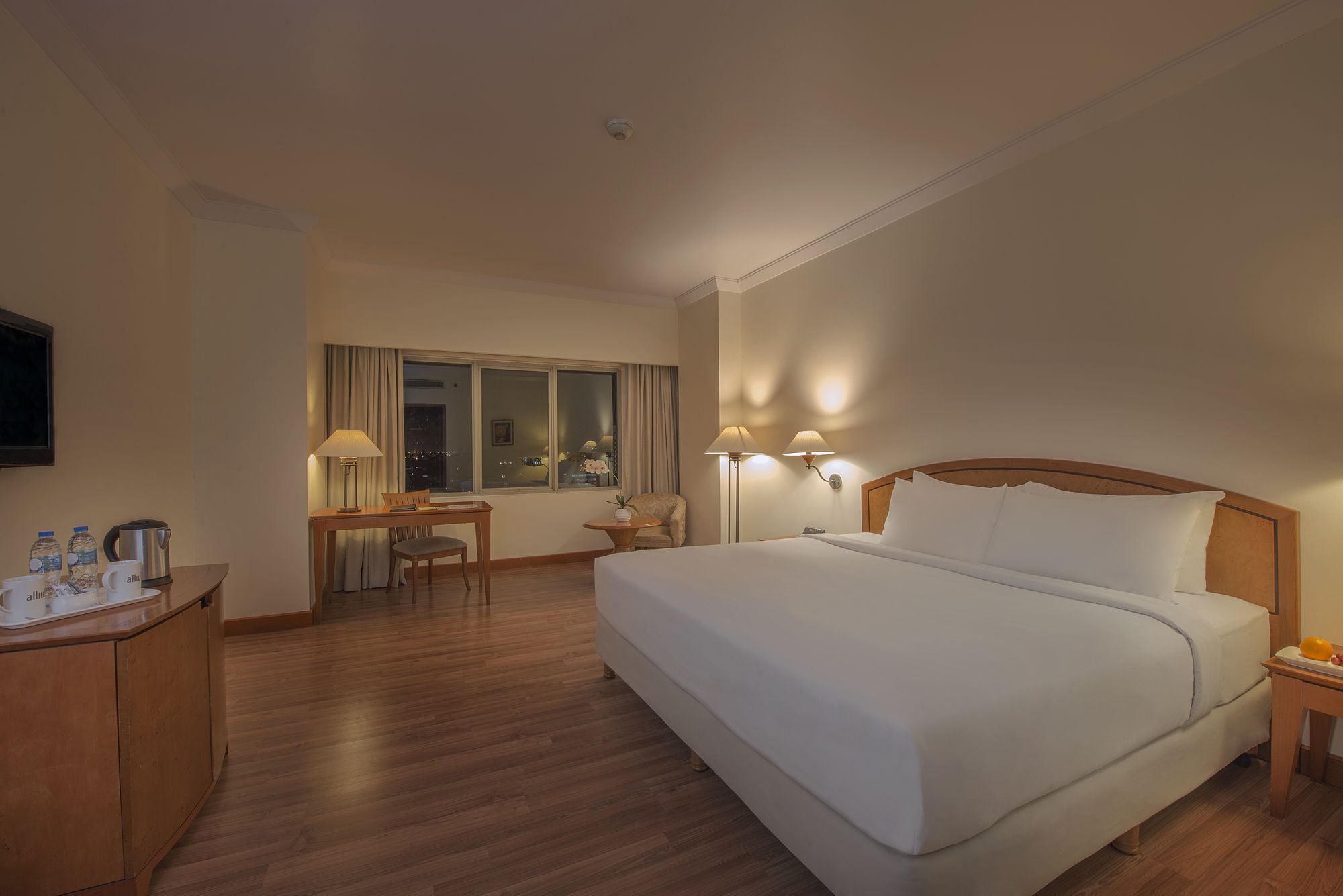 A hotel room with a large bed, a desk with a chair, a small table with a lamp, and a window with curtains.