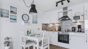 A modern kitchen with white cabinetry, a Bosch oven, and a microwave. There is a dining area with white chairs and a table set for two. - Hafenmelodie - waterfront, 1st floor, harbor view, balcony, shower room (Olpenitz)