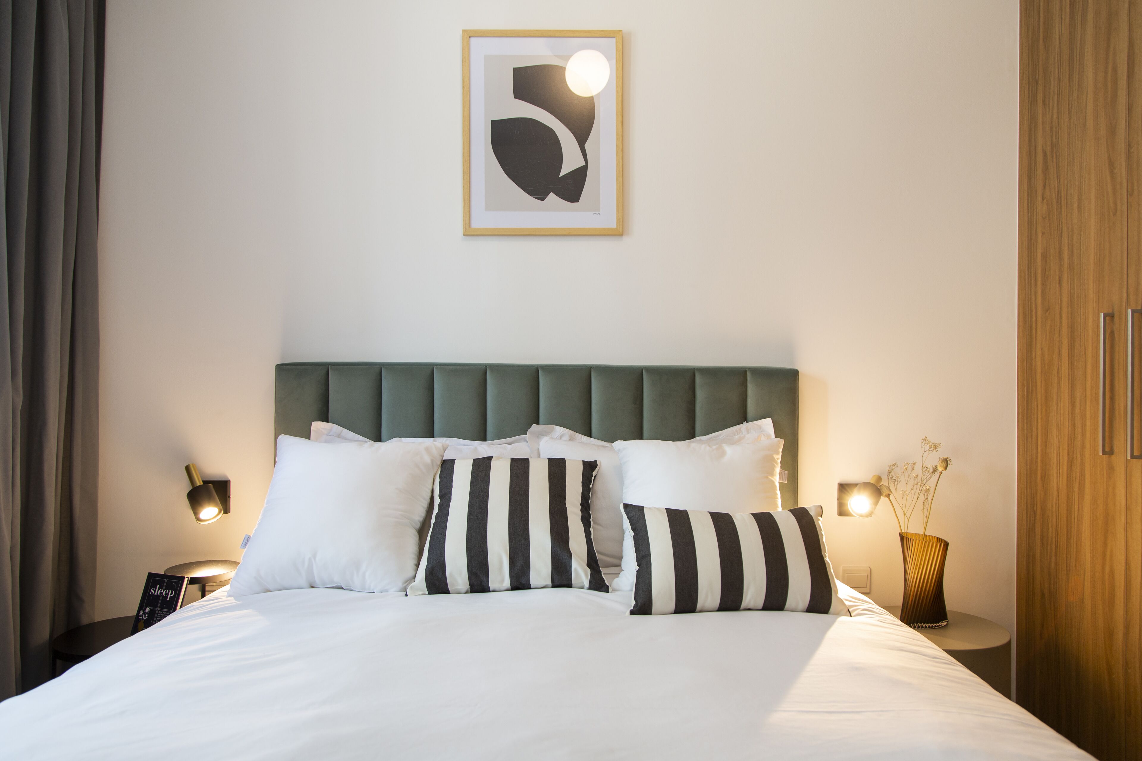 A neatly made bed with white and striped pillows, a green upholstered headboard, and a framed abstract artwork above.