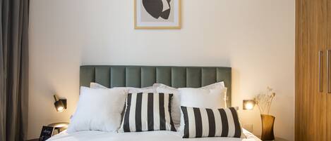 A neatly made bed with white and striped pillows, a green upholstered headboard, and a framed abstract artwork above.