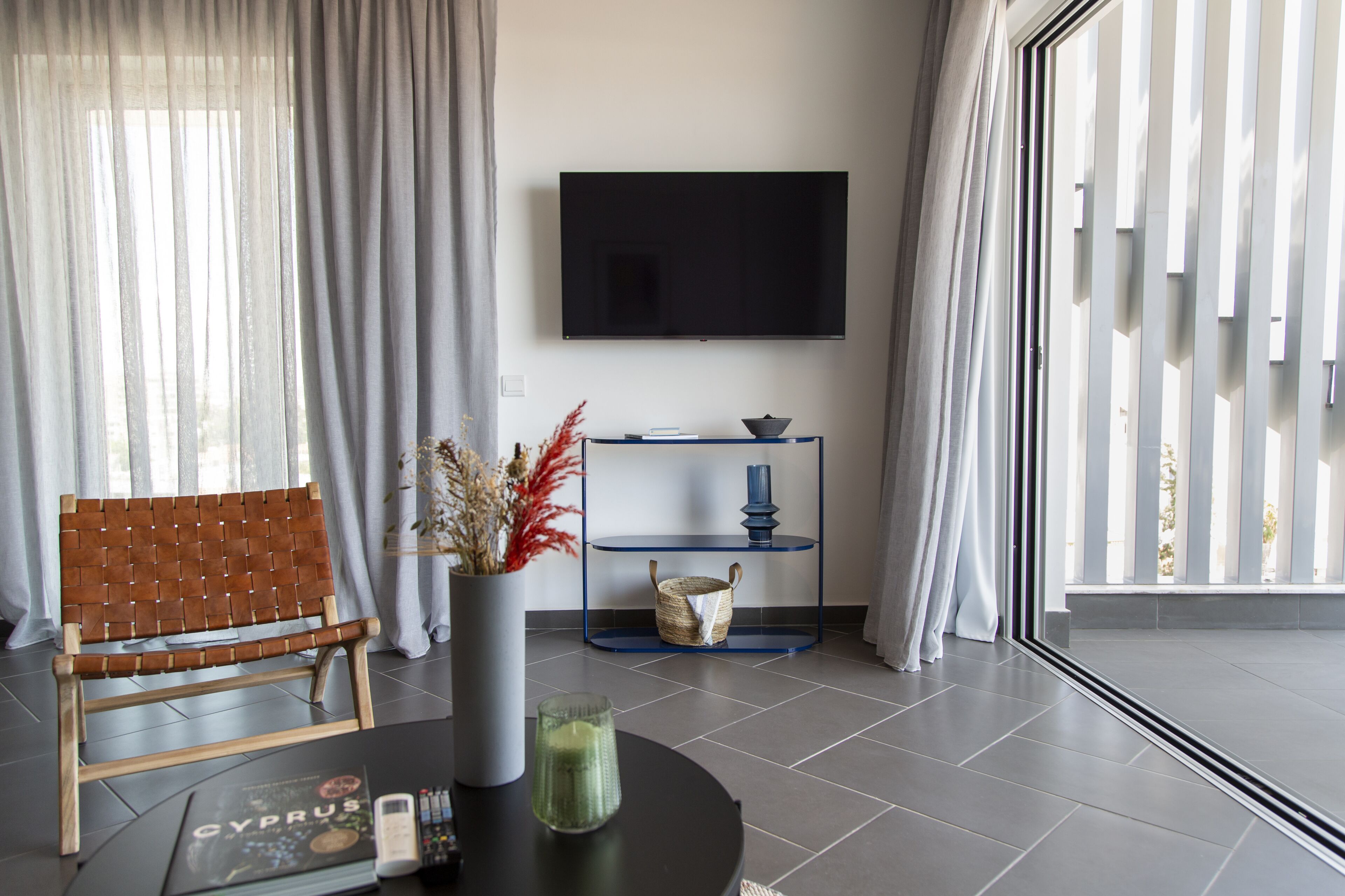 Un soggiorno moderno con una poltrona in pelle, una TV a schermo piatto, un tavolino consolle blu con oggetti decorativi e un'ampia finestra con tende.