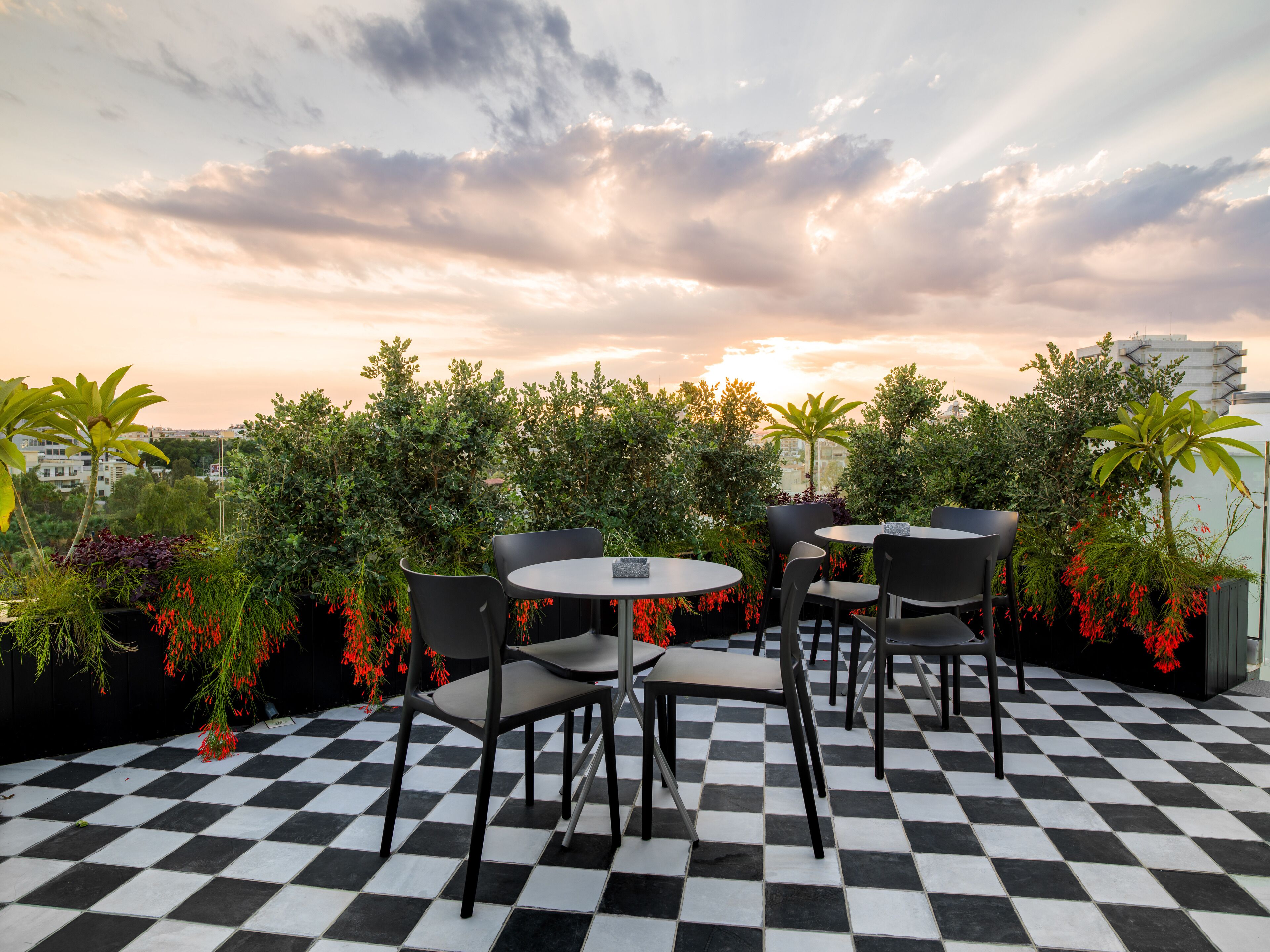 A rooftop terrace with checkered flooring, round tables, and chairs, surrounded by lush greenery and flowering plants.