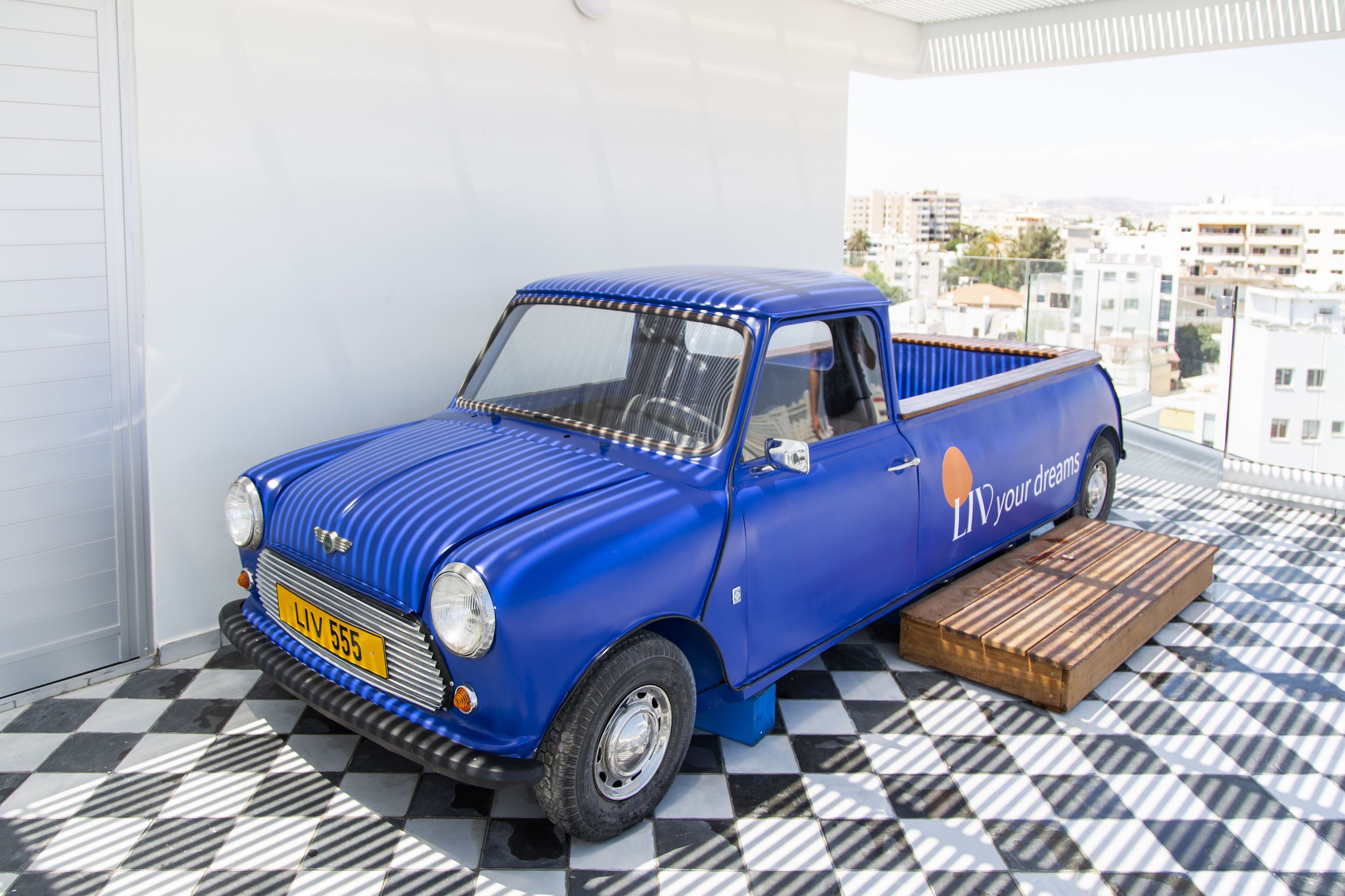 A blue vintage car parked on a balcony with a checkered floor, a city view, and a wooden platform.