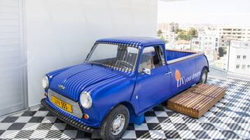 A blue vintage car parked on a balcony with a checkered floor, a city view, and a wooden platform.