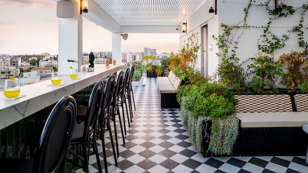 Um bar no terraço com piso xadrez, um balcão comprido com cadeiras, e vista para a paisagem urbana.