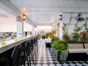 A rooftop bar with a checkered floor, a long counter with chairs, and a view of the cityscape. - LIV Urban Suites (Larnaca)