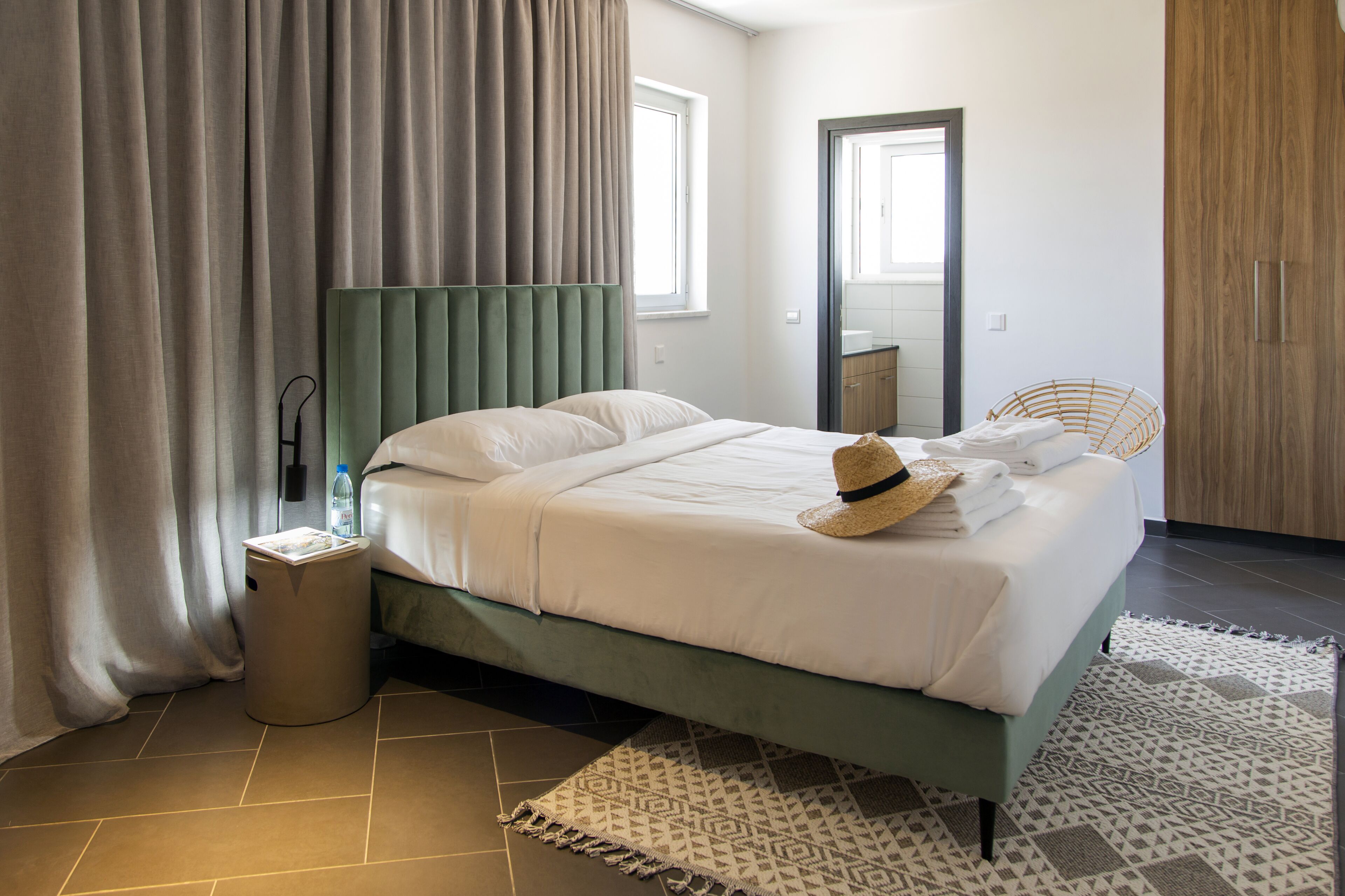 A neatly made bed with a green headboard, a small trash bin, and a woven chair.
