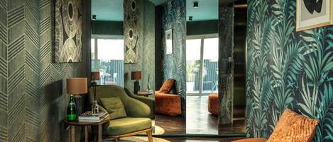 A modern living room with a green sofa, a round orange ottoman, and a patterned wall.