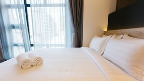 Een net opgemaakt bed met witte lakens en opgerolde handdoeken, een houten hoofdbord en uitzicht door dunne gordijnen.