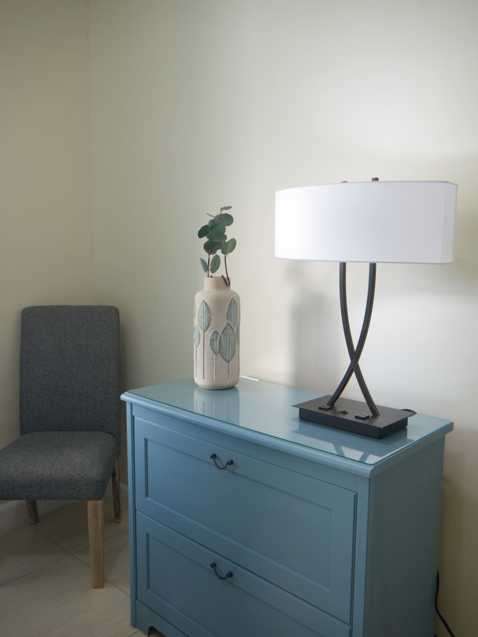 A room with a blue dresser, a grey chair, and a white lamp with a leaf design on the vase.