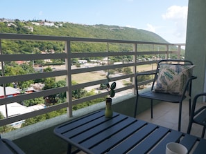 A balcony with a view of a hillside, a chair with a leaf-patterned pillow, and a small potted plant. - 2 bed 2 bath apartment exactly across from the National Stadium/Arena  (Kingston)