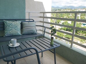 A balcony with a metal table and bench, a white railing, and a view of a cityscape with greenery and mountains in the distance.