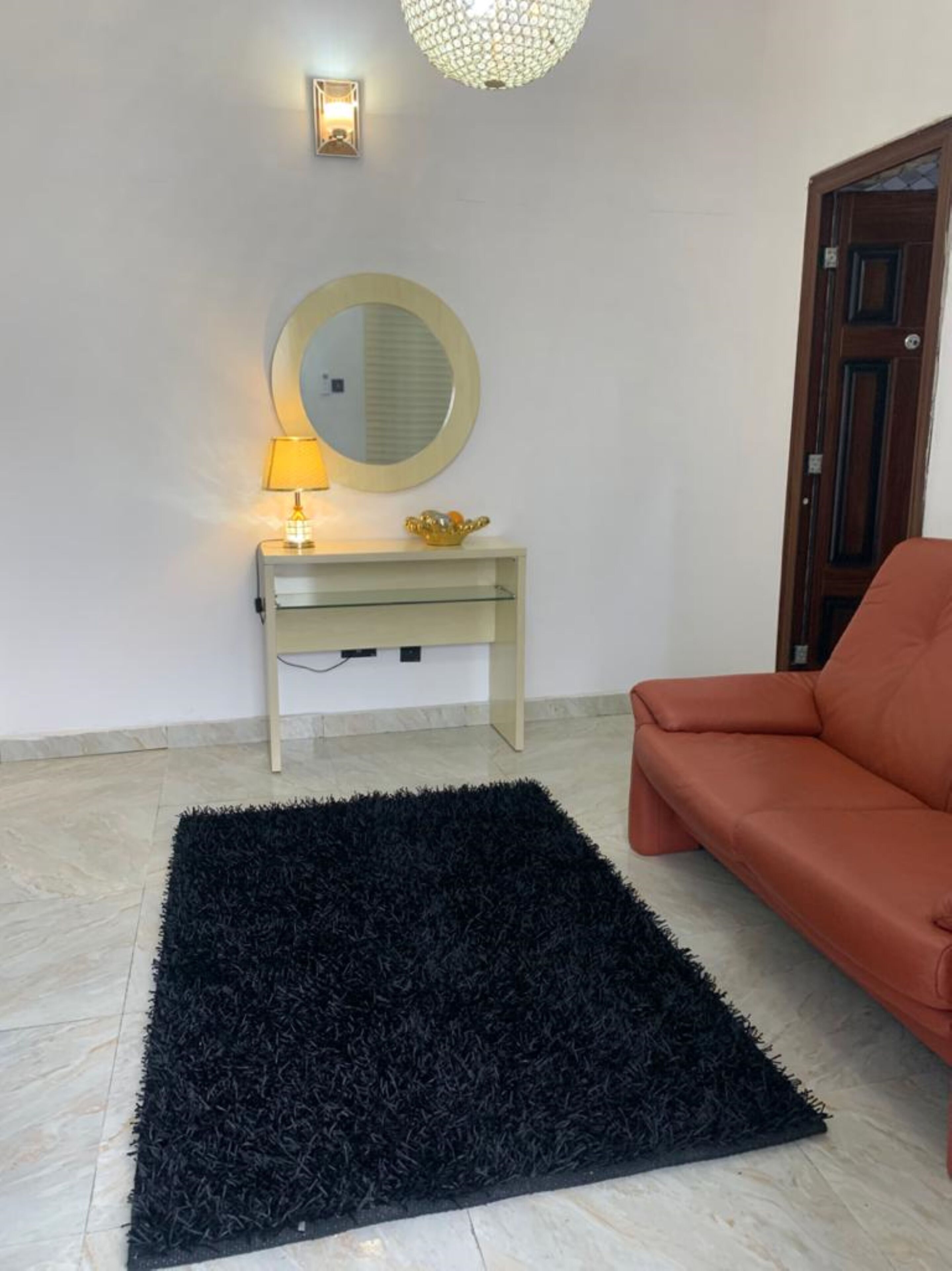 A room with a black rug, a round mirror, a small table with a lamp, and a brown leather sofa.