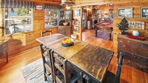A cozy cabin interior with a wooden dining table, chairs, a fireplace, a pool table, and a sign that reads "THE CABIN IS MY HAPPY PLACE."
