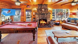 A cozy lodge interior with a pool table, leather seating, a fireplace, and a large window overlooking a forested area.