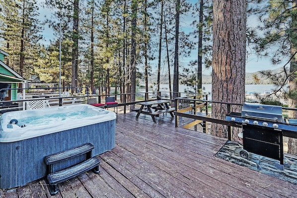 Una terraza de madera con jacuzzi, mesas de picnic y una parrilla asadora con vistas a un lago.