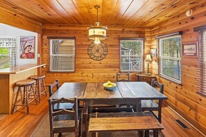 L’intérieur d’un chalet en bois, avec une table à manger, des chaises, une horloge et une fenêtre avec des stores.