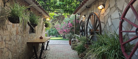 A stone-walled garden with a wooden table and benches, potted plants, and a stone pathway leading to a lush garden area.
