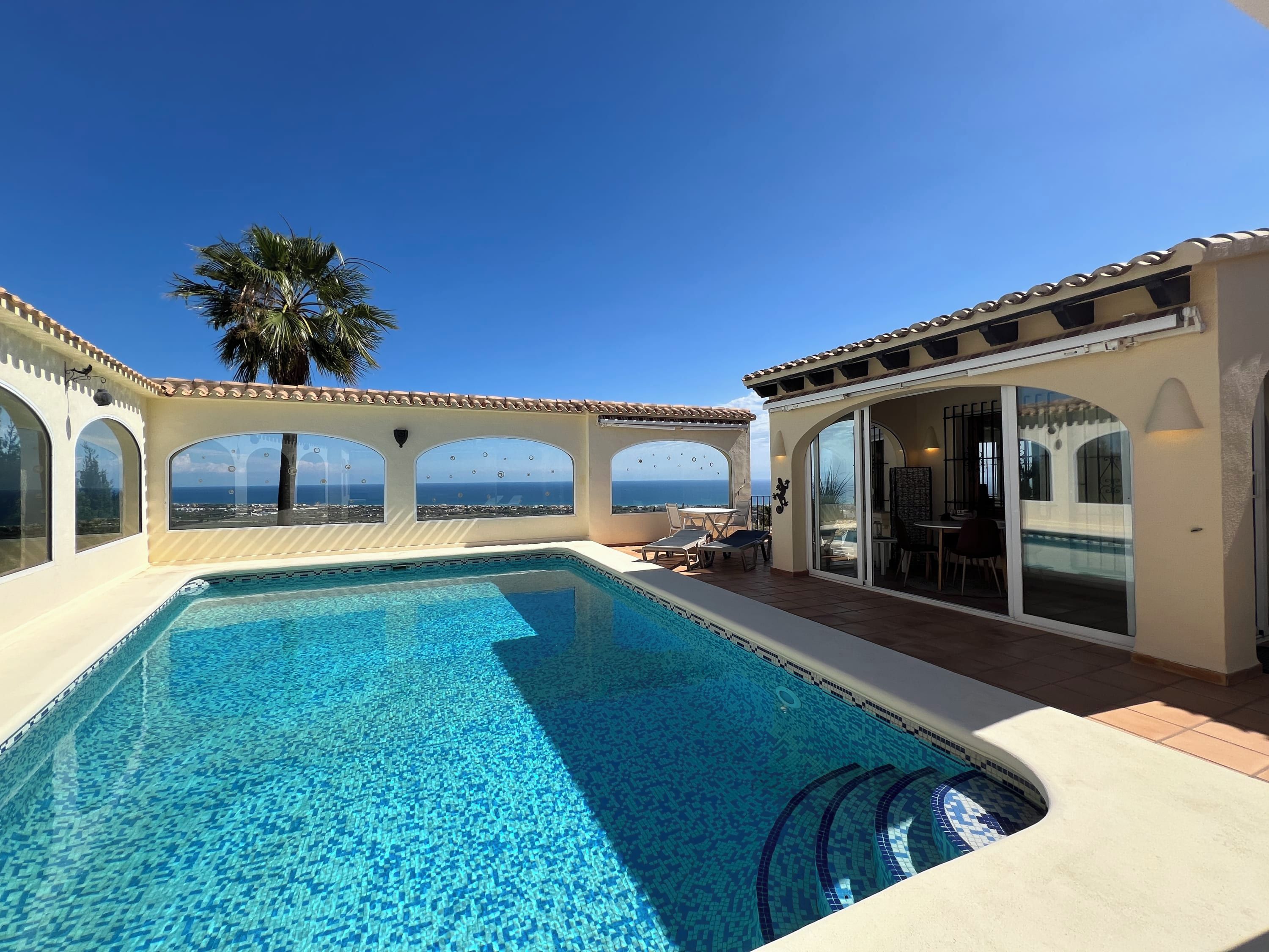 A swimming pool with a view of the ocean, a palm tree, and a covered patio area.