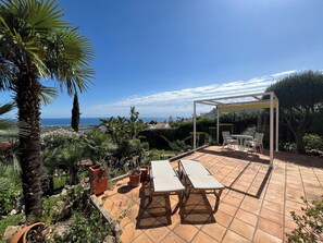 A patio with a table and chairs, surrounded by greenery and a view of the sea.