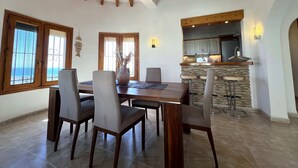 A dining area with a wooden table and chairs, a kitchen with stone accents, and large windows with decorative grills. - T-0121, Canarios, Fantastic villa with panoramic sea views (Denia)