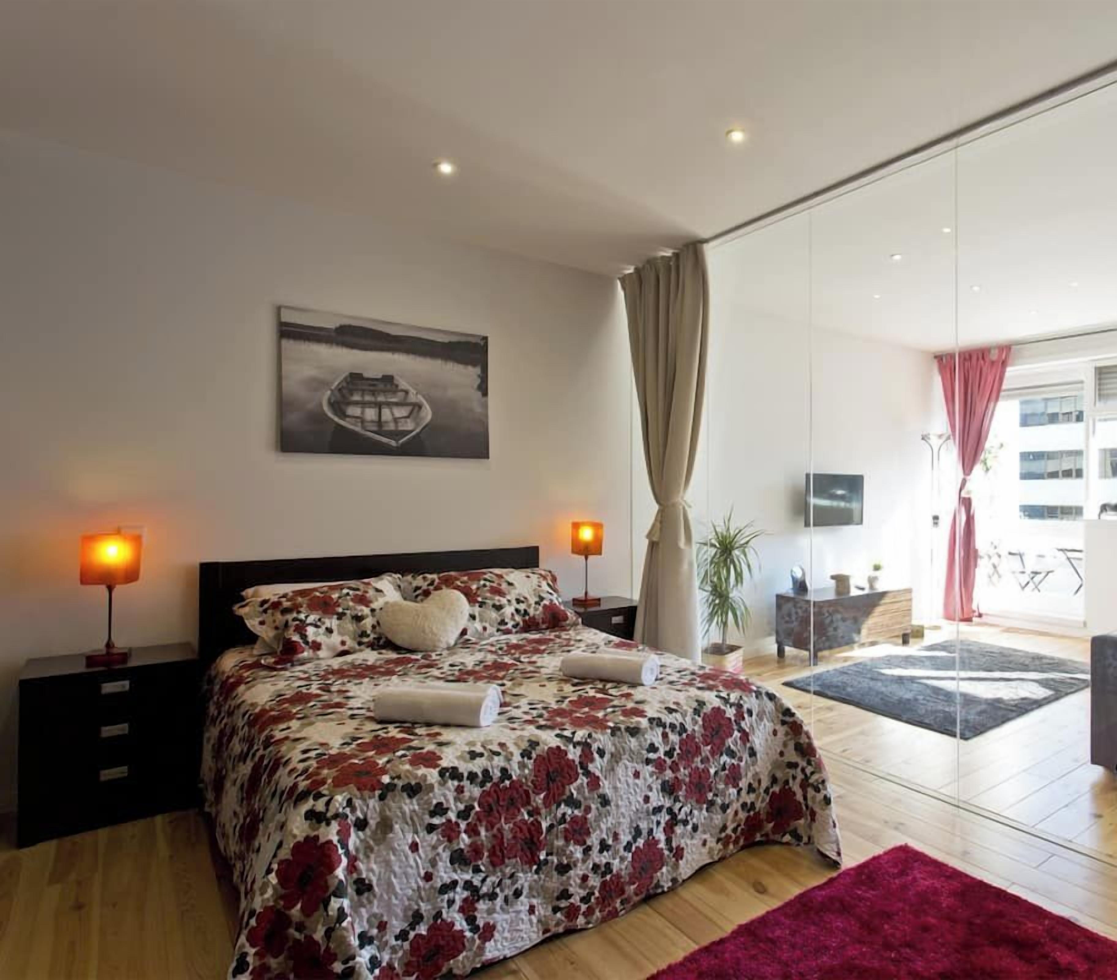 Une chambre moderne avec un grand lit, une table de chevet avec une lampe, une fenêtre avec des rideaux et une vue sur l’extérieur.