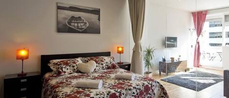 Un dormitorio moderno con una cama grande, una mesita de noche con lámpara, una ventana con cortinas y vistas al exterior.