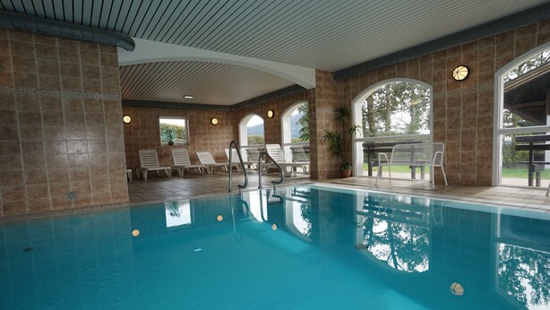Una piscina interior con agua cristalina, rodeada de sillones de descanso y una zona cubierta con vistas a zonas verdes.
