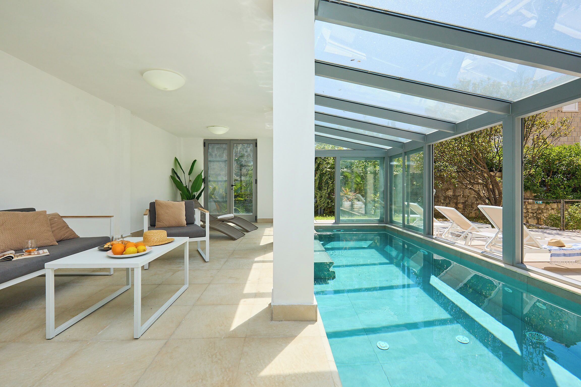 Uma área interna de piscina moderna com teto de vidro, um sofá, uma mesa com frutas e vista para o exterior.
