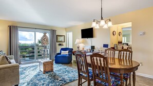 A spacious living room with a large sliding glass door leading to a balcony, a dining area with a round table and wooden chairs, and a kitchen visible through an open doorway. - Private balcony overlooking the beach! (Tybee Island)