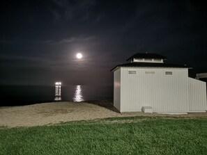 Un bâtiment blanc avec un toit noir, portant l’inscription « BLUE FISH », situé près d’une plage la nuit, avec une lune visible et des reflets sur l’eau.