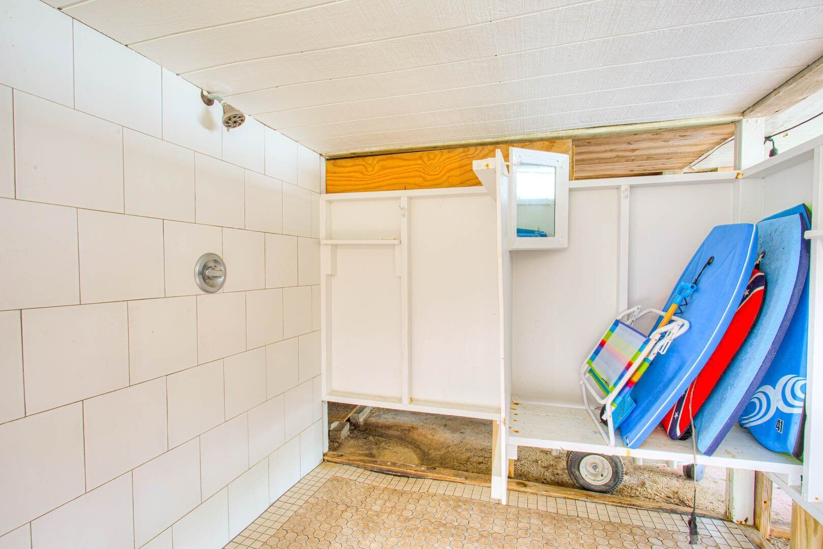 A small, enclosed room with white tiled walls, a wooden ceiling, and a white shelf with surfboards.