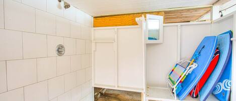 A small, enclosed room with white tiled walls, a wooden ceiling, and a white shelf with surfboards.