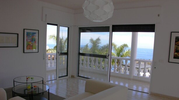 A spacious living room with a white sofa, a glass coffee table, and a view of the sea through large sliding doors.
