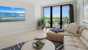 A spacious living room with a large flat-screen TV, a beige sofa, a round coffee table, and a balcony with outdoor seating. - Beachfront 2BR/2BA condo with heated pool, WiFi, and Marco Island views. (Marco Island)