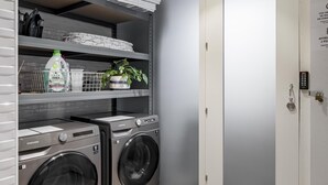 Un lavadero con dos lavadoras de carga frontal, un estante con productos de limpieza y una puerta con cerradura.