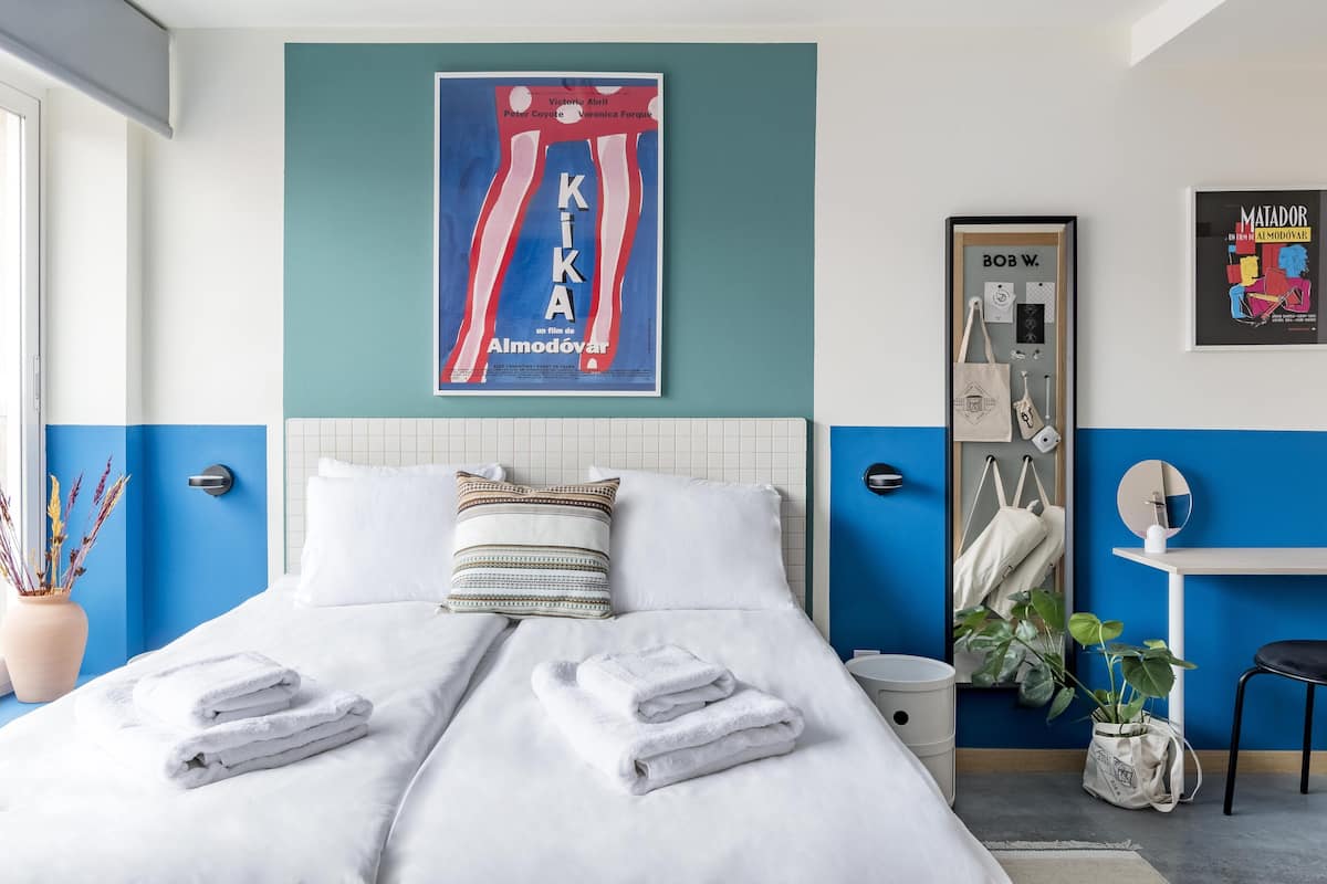 Una cama bien hecha con toallas dobladas, un cartel de película enmarcado, un espejo, una planta y una silla en una habitación moderna con paredes azules y blancas.