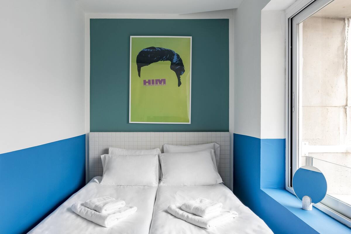 Habitación de hotel con cama, dos almohadas, una toalla, una ventana y una obra de arte colgada en la pared.