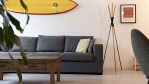 Ein Wohnzimmer mit grauem Sofa, einem Holztisch, einem Surfbrett an der Wand und einem Stativ mit einem gerahmten Bild.