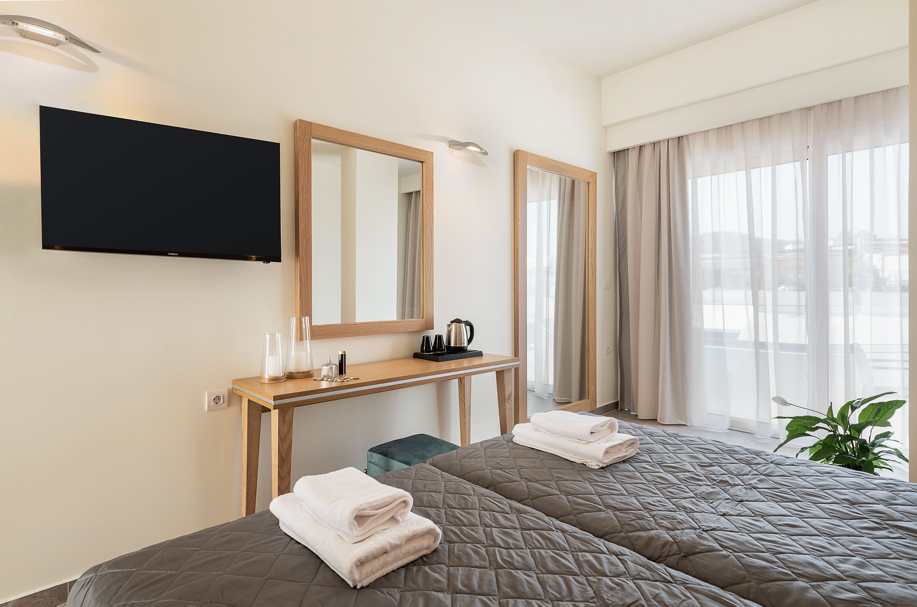 A hotel room with two beds, a TV, a desk with a mirror, a kettle, and a plant.