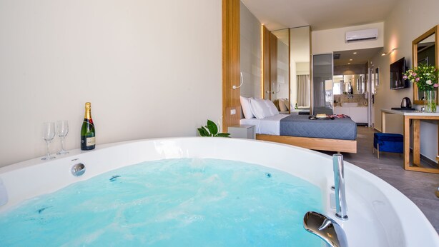 A modern hotel room with a large bathtub, a bed with a gray quilted mattress, a wooden bedside table, and a small desk with a chair. - Elia Platanias (Platanias)