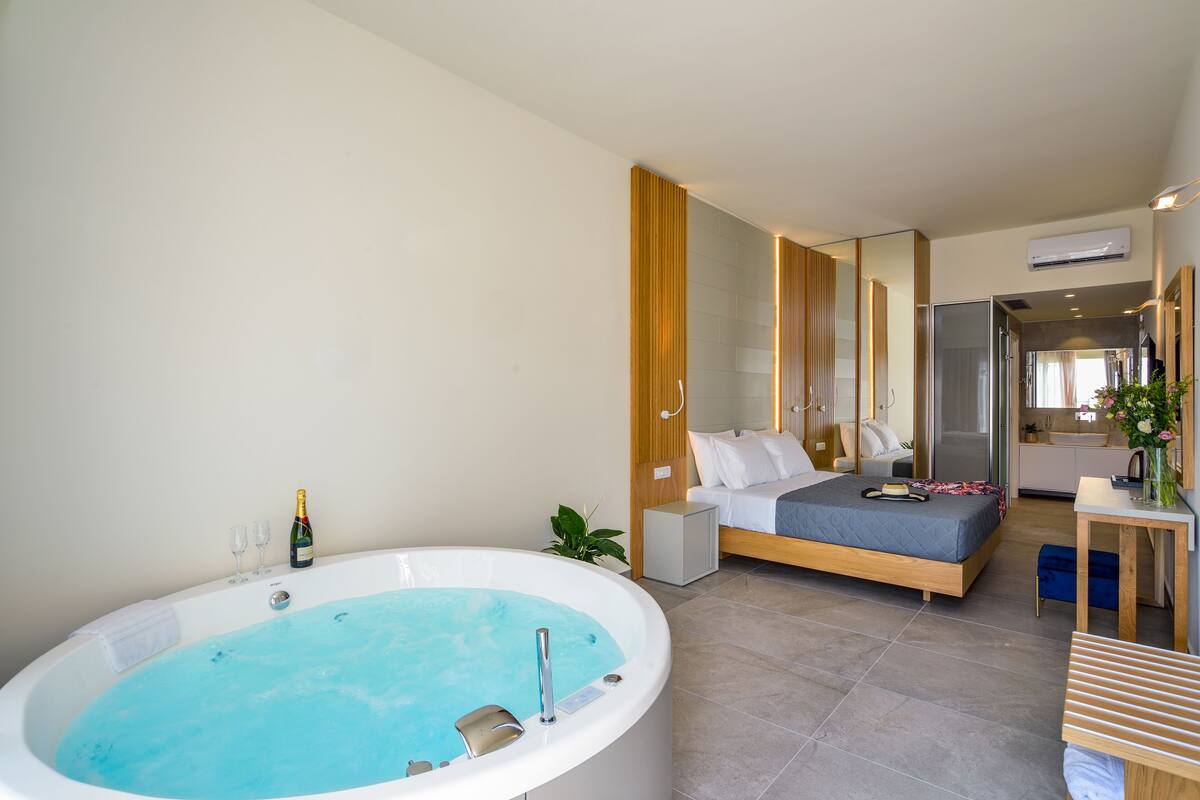 A modern bedroom with a large bathtub, a bed, a bedside table, and a mirror.