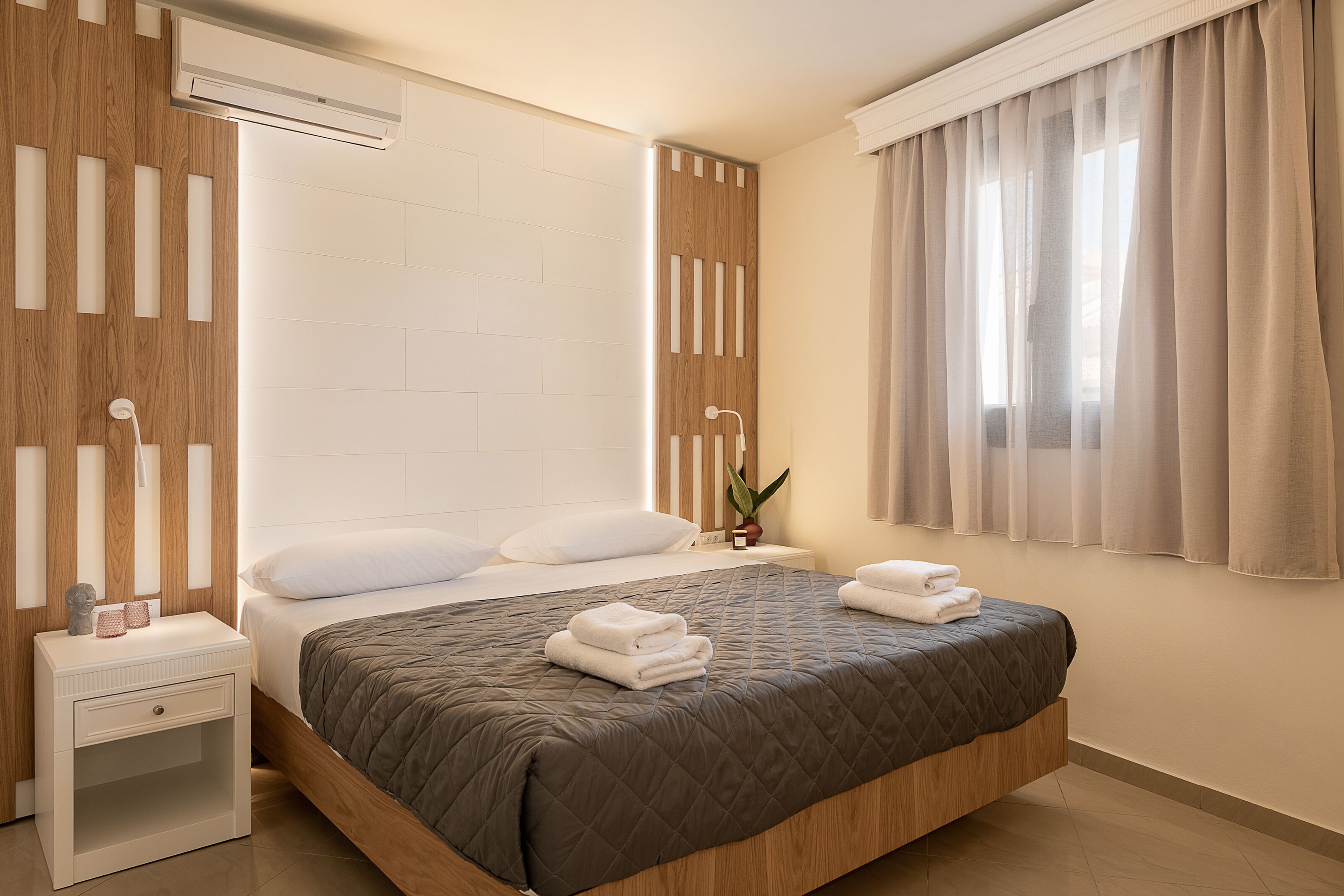 A modern bedroom with a large bed, a nightstand, an air conditioner, a window with curtains, and a potted plant.