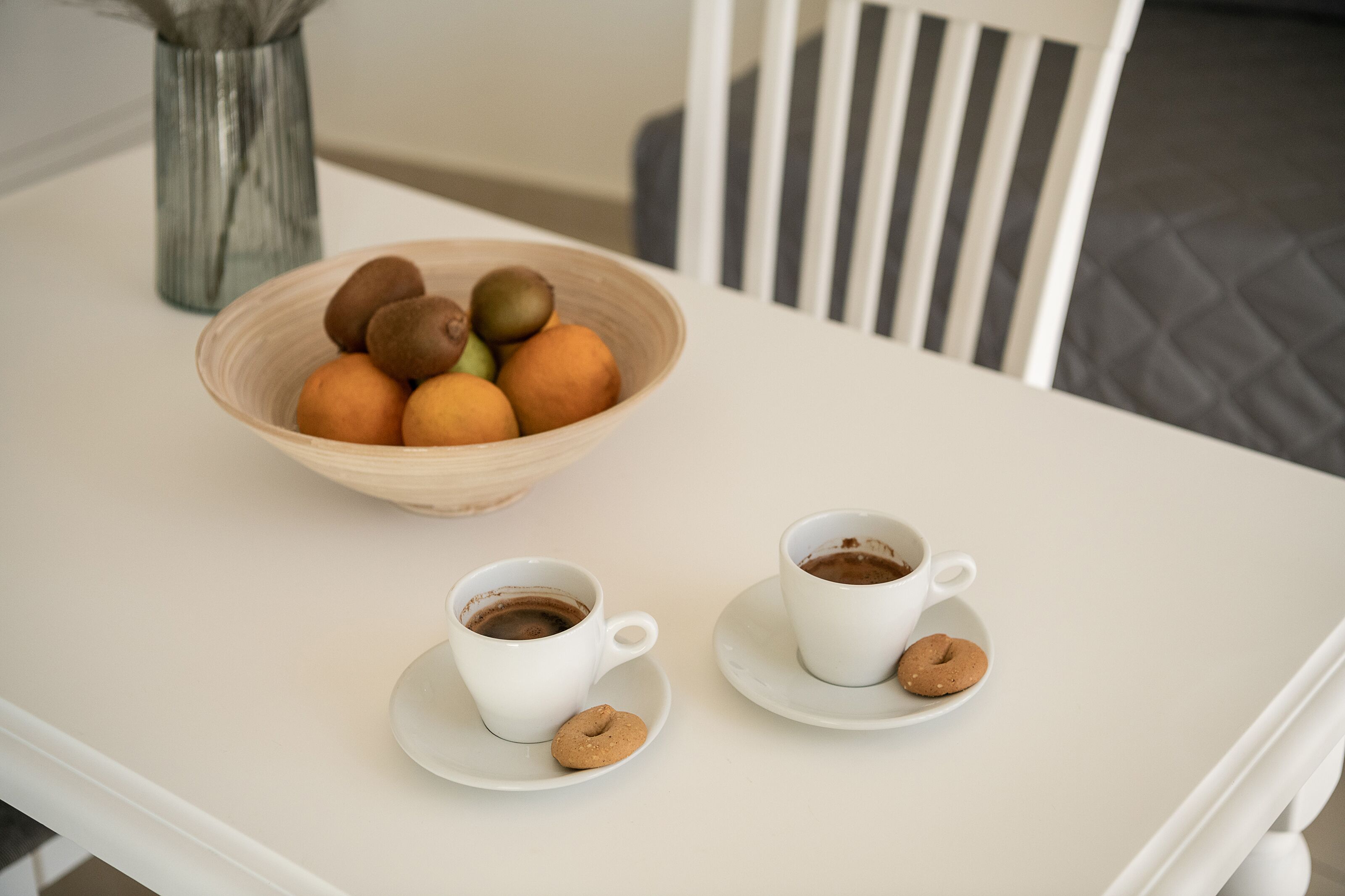 A table with a bowl of fruit, two cups of coffee, and a cookie.