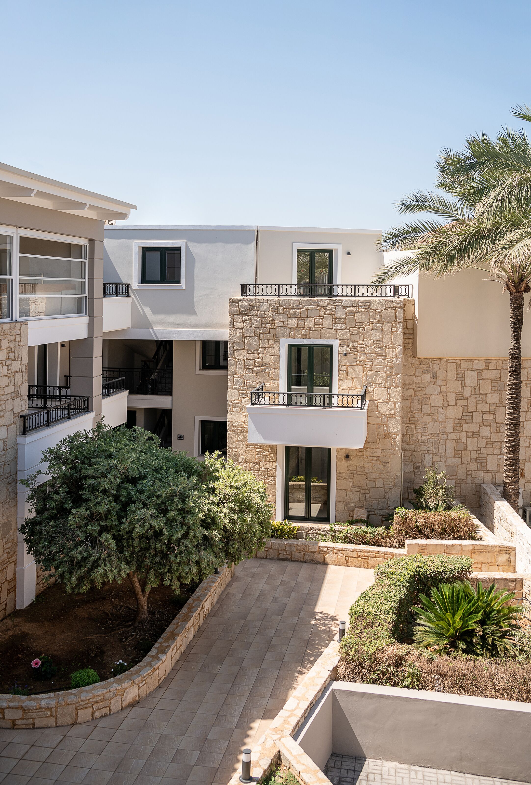 A multi-story building with balconies, a stone facade, and a landscaped courtyard with a palm tree and shrubs.