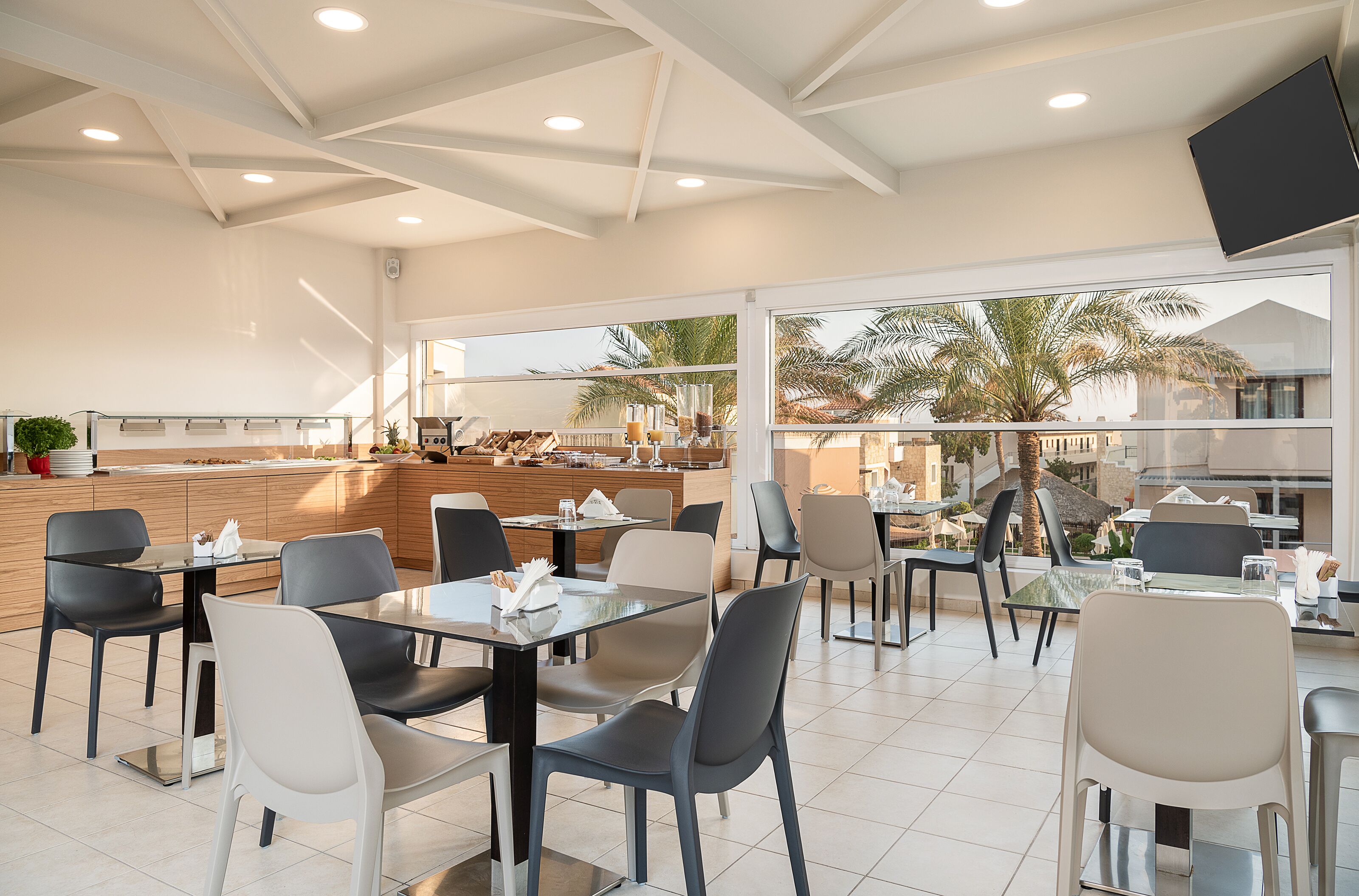 A modern restaurant with a spacious layout, large windows offering views of palm trees and a pool, and a central island with a bar area.