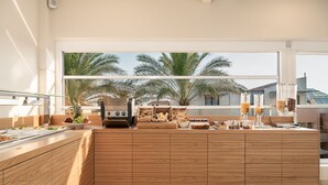 A modern kitchen with a large island, wooden cabinetry, and a view of palm trees through large windows. - Elia Platanias (Platanias)