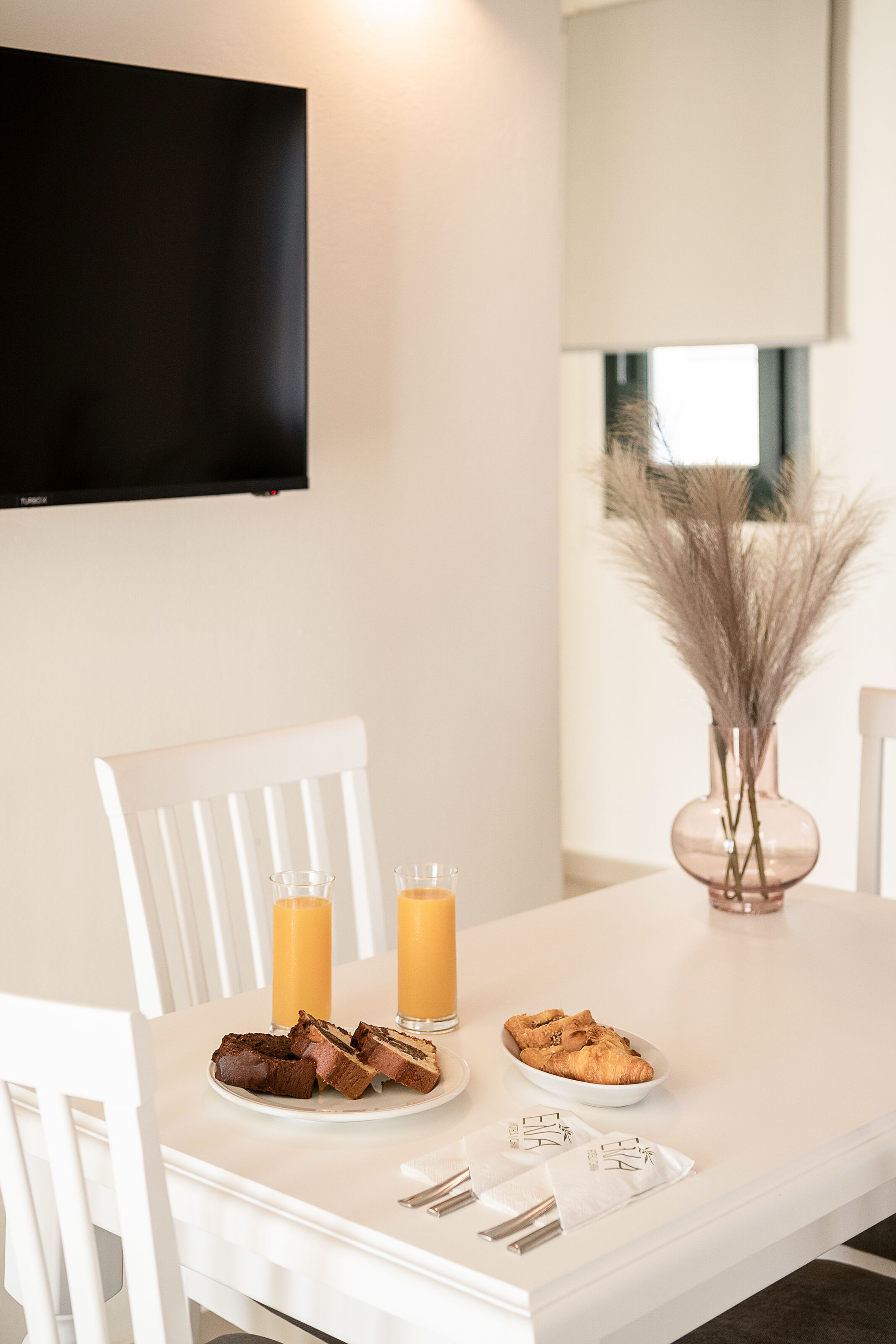Une table à manger avec une assiette de nourriture, deux verres de jus d’orange et un vase contenant des plantes séchées.