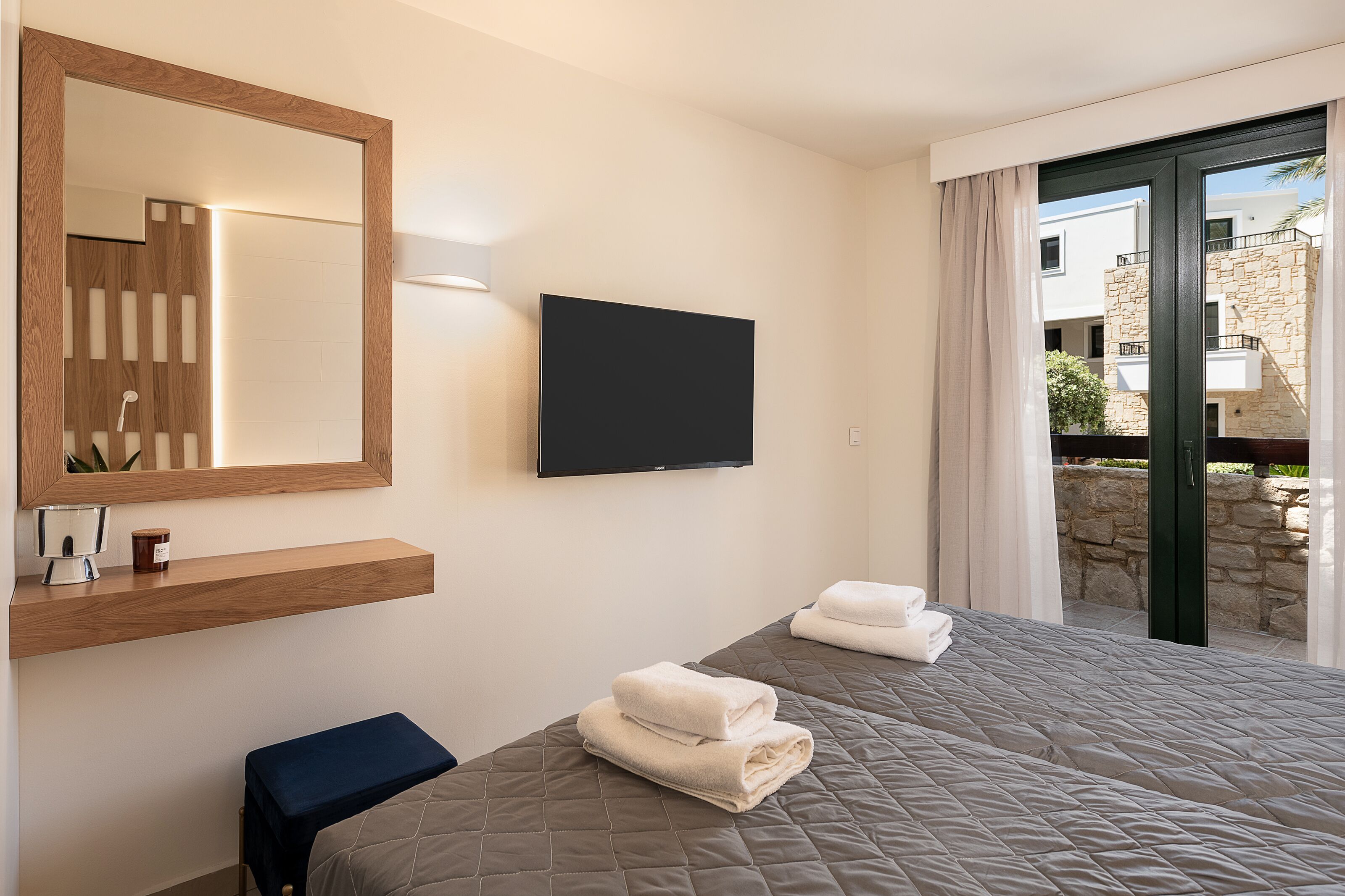 A modern hotel room with a large bed, a flat-screen TV, a wooden shelf with a candle, and a view of the outside balcony.
