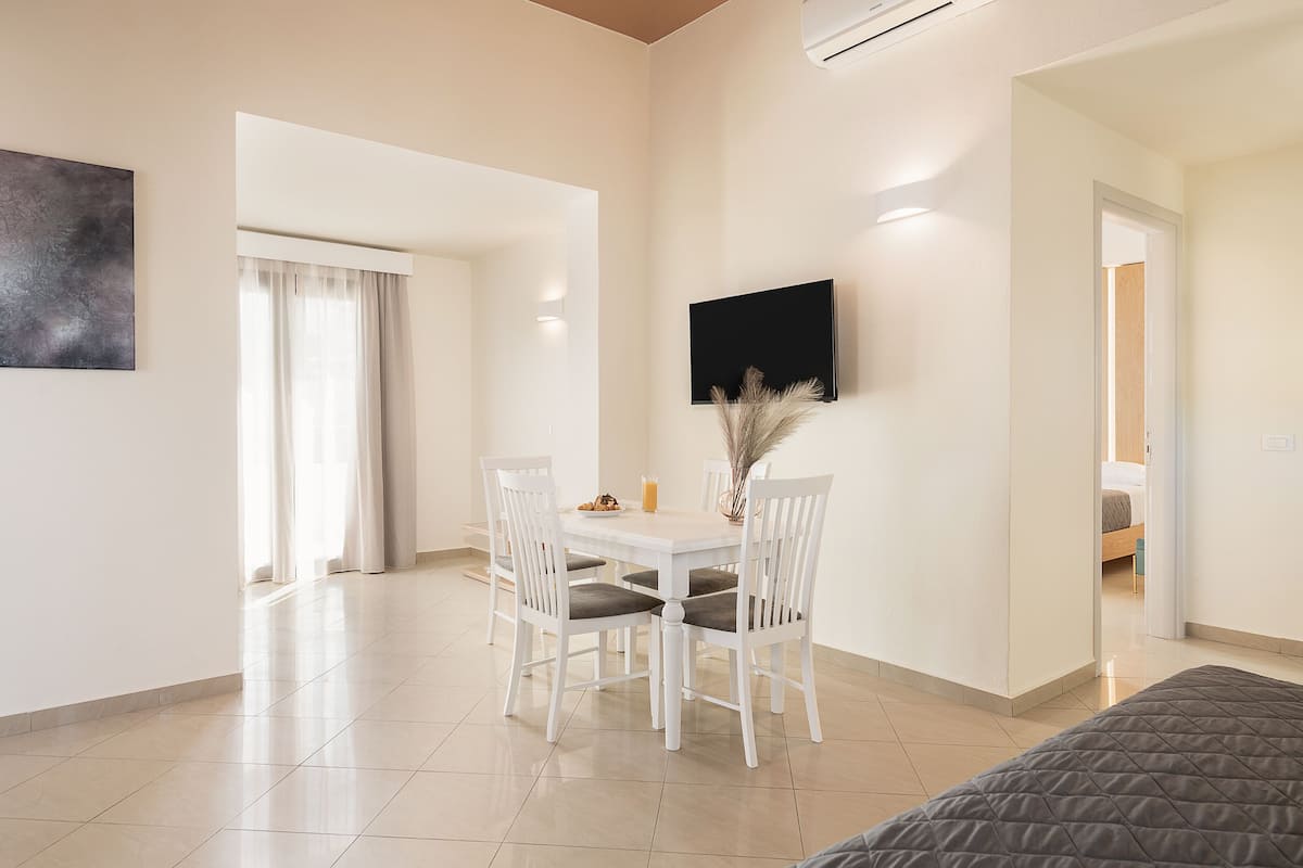 A modern hotel room with a bed, a dining table with chairs, a flat-screen TV mounted on the wall, and a large window with curtains.